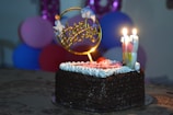 A chocolate cake is topped with white frosting, a pink rose decoration, and several candles. A decorative topper reads 'Happy Birthday' with small cartoon figures. In the background, a cluster of colorful balloons in shades of blue, pink, and purple is visible.