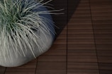 A modern ceramic planter filled with long, thin blades of grass. The planter is placed on a textured, wooden deck, with shadows cast across the surface.