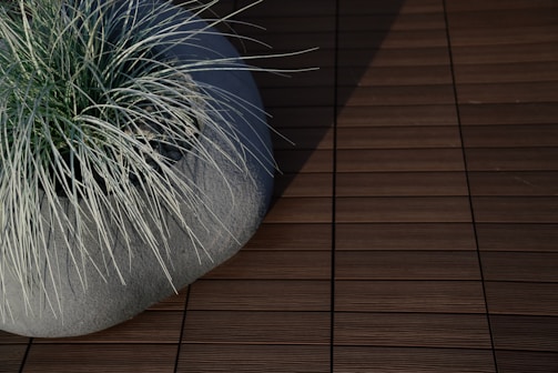 A modern ceramic planter filled with long, thin blades of grass. The planter is placed on a textured, wooden deck, with shadows cast across the surface.