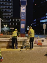 Two construction workers in high-visibility vests and helmets are performing maintenance or construction work on a street at night. They are positioned on either side of a blue and red signpost with labels, including A2. Various construction materials and tools are scattered on the pavement near them, and a building and some trees are visible in the background.