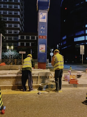 Two construction workers in high-visibility vests and helmets are performing maintenance or construction work on a street at night. They are positioned on either side of a blue and red signpost with labels, including A2. Various construction materials and tools are scattered on the pavement near them, and a building and some trees are visible in the background.
