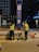 Two construction workers in high-visibility vests and helmets are performing maintenance or construction work on a street at night. They are positioned on either side of a blue and red signpost with labels, including A2. Various construction materials and tools are scattered on the pavement near them, and a building and some trees are visible in the background.