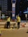 Two construction workers in high-visibility vests and helmets are performing maintenance or construction work on a street at night. They are positioned on either side of a blue and red signpost with labels, including A2. Various construction materials and tools are scattered on the pavement near them, and a building and some trees are visible in the background.