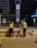 Two construction workers in high-visibility vests and helmets are performing maintenance or construction work on a street at night. They are positioned on either side of a blue and red signpost with labels, including A2. Various construction materials and tools are scattered on the pavement near them, and a building and some trees are visible in the background.