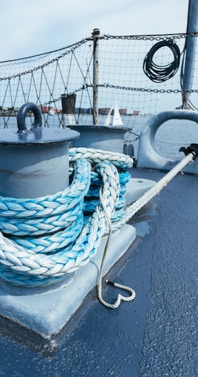 A coiled rope is neatly wrapped around a metal post on a boat deck, with a nautical background featuring a safety net, metal railings, and a view of water and distant sails.