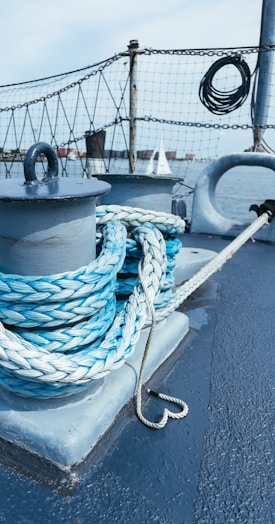 A coiled rope is neatly wrapped around a metal post on a boat deck, with a nautical background featuring a safety net, metal railings, and a view of water and distant sails.