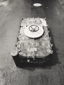 A heavy metal hatch with a circular handle is set into the ground, surrounded by a wet, textured surface. The hatch features multiple latches and bolts around its edges, indicating it is securely fastened. In the background, a circular manhole cover is visible on the asphalt.