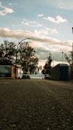A low-angle view of a street with a car parked on the left and portable toilets lined up on the right side. Trees with sparse leaves are seen along the street, and in the background, a cloudy sky is visible.