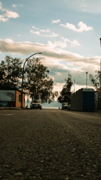 A low-angle view of a street with a car parked on the left and portable toilets lined up on the right side. Trees with sparse leaves are seen along the street, and in the background, a cloudy sky is visible.
