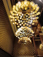A large, intricate chandelier composed of numerous circular light fixtures suspended from the ceiling. The lights are arranged in a cascading pattern, illuminating the space with a warm, golden glow. The surrounding walls feature a textured pattern that complements the chandelier's design, adding to the overall opulence and elegance of the setting.