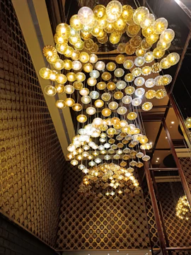 A large, intricate chandelier composed of numerous circular light fixtures suspended from the ceiling. The lights are arranged in a cascading pattern, illuminating the space with a warm, golden glow. The surrounding walls feature a textured pattern that complements the chandelier's design, adding to the overall opulence and elegance of the setting.