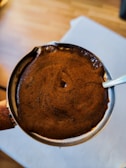 A close-up view of a freshly brewed cup of coffee with a rich layer of frothy crema on top. The cup appears to be metallic with a small handle partially visible. The background is slightly blurred, with wooden flooring and a white rectangular area possibly representing a table or cloth.