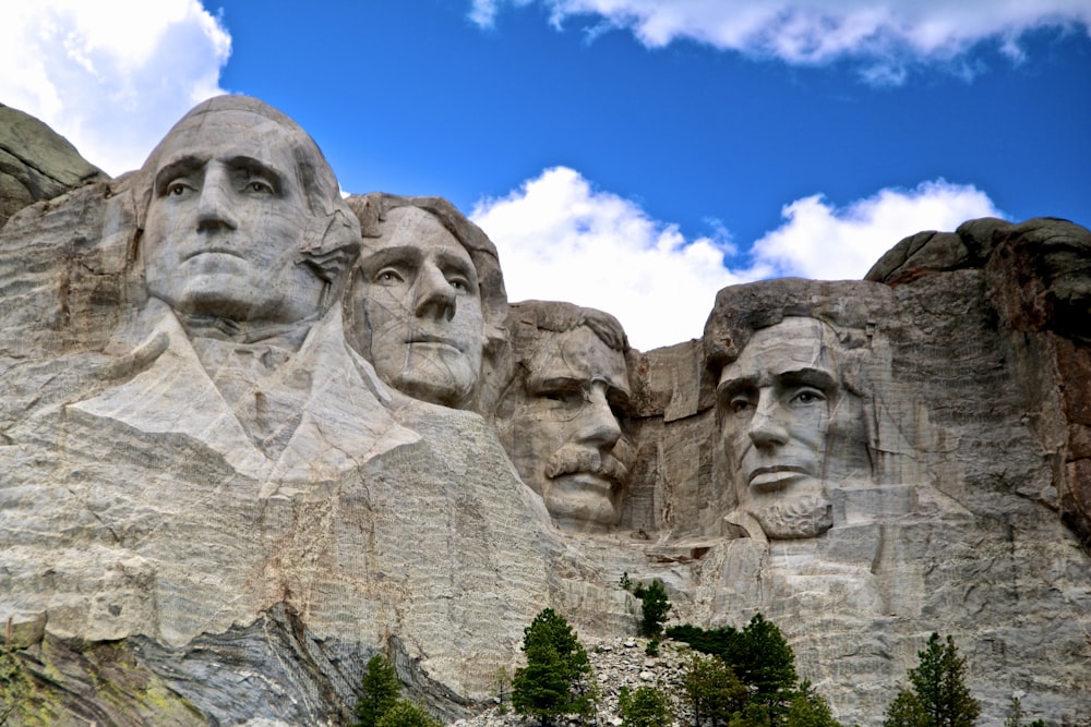 un grupo de caras talladas en una roca con el Monumento Nacional del Monte Rushmore en el fondo