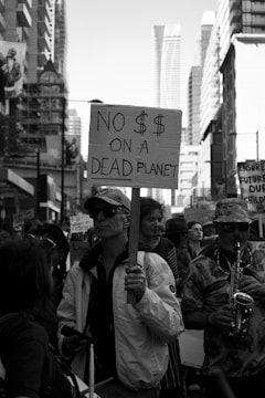 A monochrome image of a protest march in an urban setting, featuring a central figure holding a placard that reads 'No $$ on a Dead Planet'. A diverse group of people walks alongside, displaying various signs and messages. Tall buildings and city structures form the background, while one participant plays a saxophone.