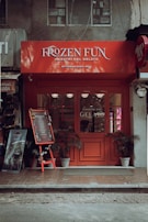 A quaint gelato shop with a vibrant red facade and signage reading 'Frozen Fun Maestri Del Gelato'. Decorative chalkboard stand displaying a menu, surrounded by potted plants. The setting appears cozy with a hint of vintage charm.
