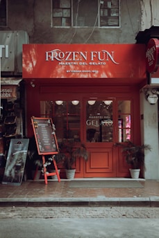 A quaint gelato shop with a vibrant red facade and signage reading 'Frozen Fun Maestri Del Gelato'. Decorative chalkboard stand displaying a menu, surrounded by potted plants. The setting appears cozy with a hint of vintage charm.