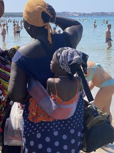 A crowded beach scene with many people in swimwear enjoying the water. In the foreground, a woman is carrying a child on her back using a colorful wrap. Both are wearing headscarves, with the child dressed in a patterned orange garment. The background shows clear blue water and distant buildings across the bay.