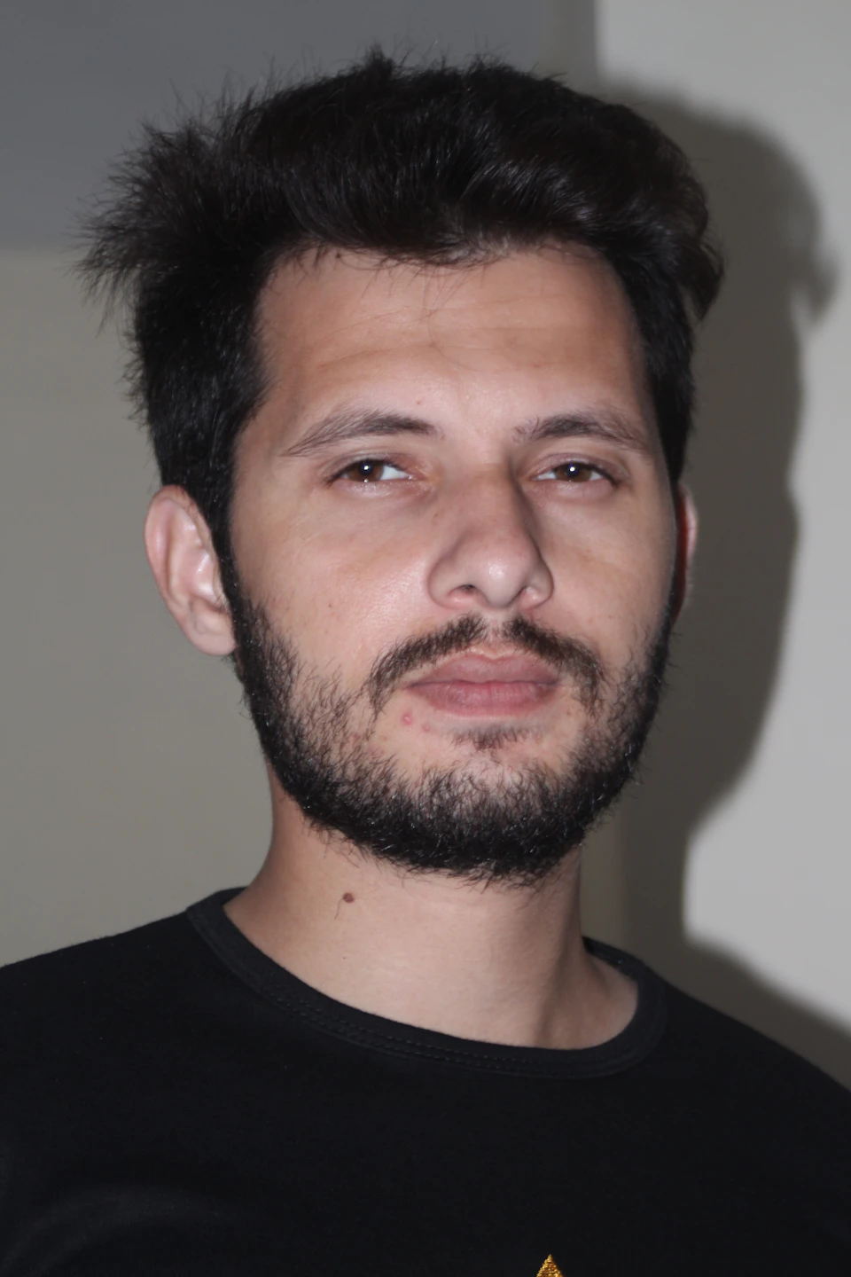 The image shows a man with short dark hair and a beard, wearing a black t-shirt. He is looking directly at the camera with an expressionless face. The background appears to be a plain white wall, which contrasts with the man's dark attire and adds depth to the scene.