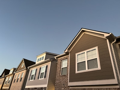 A row of modern suburban houses stretches across the image, featuring a blend of brick and siding exteriors. The architecture includes gable roofs and multiple window styles, illuminated by soft, early evening light.