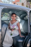 A woman in a wedding dress is sitting in the back seat of a vintage car, holding a bouquet of purple, pink, and white flowers. The car's interior features leather seating and a softly lit ambiance. Outside the window, there are some architectural elements and greenery, suggesting an outdoor setting.
