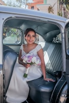 A woman in a wedding dress is sitting in the back seat of a vintage car, holding a bouquet of purple, pink, and white flowers. The car's interior features leather seating and a softly lit ambiance. Outside the window, there are some architectural elements and greenery, suggesting an outdoor setting.