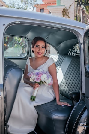 A woman in a wedding dress is sitting in the back seat of a vintage car, holding a bouquet of purple, pink, and white flowers. The car's interior features leather seating and a softly lit ambiance. Outside the window, there are some architectural elements and greenery, suggesting an outdoor setting.