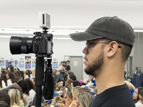 A person wearing a dark cap and glasses is holding a camera mounted on a monopod. They appear focused, surrounded by a crowd of seated individuals in a brightly lit indoor setting.