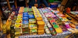 A display of neatly organized tins of preserved fish, each with distinct and colorful packaging. The tins are arranged on a decorated tablecloth, and there are people standing behind the table. Various brand names and vibrant designs are visible, suggesting a market or exhibition setting.