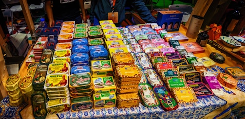 A display of neatly organized tins of preserved fish, each with distinct and colorful packaging. The tins are arranged on a decorated tablecloth, and there are people standing behind the table. Various brand names and vibrant designs are visible, suggesting a market or exhibition setting.
