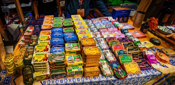 A display of neatly organized tins of preserved fish, each with distinct and colorful packaging. The tins are arranged on a decorated tablecloth, and there are people standing behind the table. Various brand names and vibrant designs are visible, suggesting a market or exhibition setting.