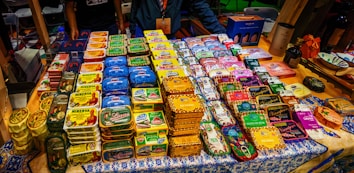 A display of neatly organized tins of preserved fish, each with distinct and colorful packaging. The tins are arranged on a decorated tablecloth, and there are people standing behind the table. Various brand names and vibrant designs are visible, suggesting a market or exhibition setting.