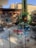 A variety of colorful metal garden sculptures are displayed outdoors on a paved surface. They range in design, including floral patterns and abstract shapes. A building with a brick facade and tall trees add dimension to the background.