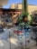 A variety of colorful metal garden sculptures are displayed outdoors on a paved surface. They range in design, including floral patterns and abstract shapes. A building with a brick facade and tall trees add dimension to the background.