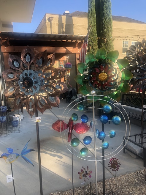 A variety of colorful metal garden sculptures are displayed outdoors on a paved surface. They range in design, including floral patterns and abstract shapes. A building with a brick facade and tall trees add dimension to the background.