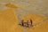 Three people are working together on a large expanse of rice being spread out to dry under the sun. Their colorful clothing contrasts with the golden hue of the grains surrounding them. The rice is laid out in geometric patterns and they appear to be using tools to organize the spread of the grains.