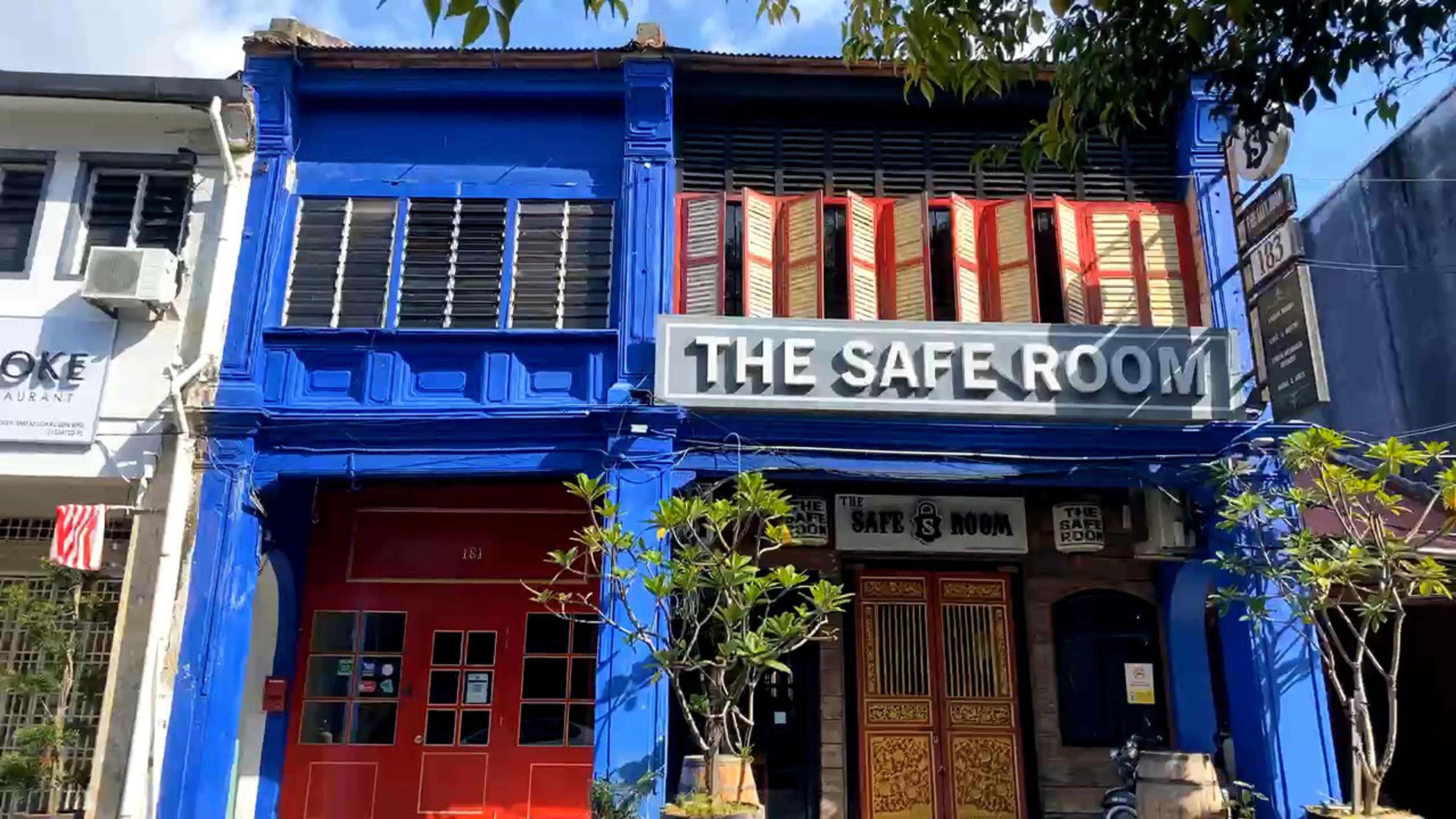 A blue heritage shophouse with red shutters and a hand-painted 'The Safe Room' sign, tropical plants out front.
