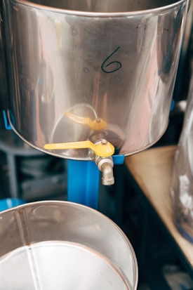 A metallic container marked with the number 6 features a yellow spigot attached near the bottom. The container is polished, reflecting some light, and sits atop a blue framework. Below it, a similar container can be seen, ready to collect any liquid dispensed from the upper one.