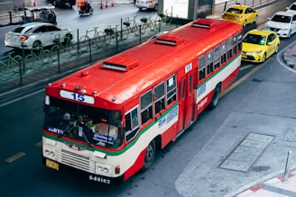 A red and white public bus marked with the number 15 is traveling on a city street. The street is shared with several other vehicles, including taxis and cars. There are green bike racks along the sidewalk, and orange traffic cones can be seen in the background. The setting appears to be urban, with a few people visible within the bus.