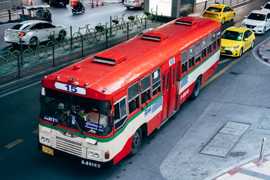 A red and white public bus marked with the number 15 is traveling on a city street. The street is shared with several other vehicles, including taxis and cars. There are green bike racks along the sidewalk, and orange traffic cones can be seen in the background. The setting appears to be urban, with a few people visible within the bus.