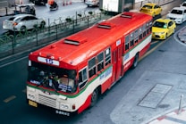 A red and white public bus marked with the number 15 is traveling on a city street. The street is shared with several other vehicles, including taxis and cars. There are green bike racks along the sidewalk, and orange traffic cones can be seen in the background. The setting appears to be urban, with a few people visible within the bus.