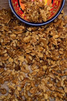 A colorful ceramic bowl is partially filled with shelled walnuts, spilling over onto a wooden surface. The walnuts are light to dark brown, with intricate textures and patterns visible on their surfaces. The bowl features vibrant red, yellow, and blue patterns.