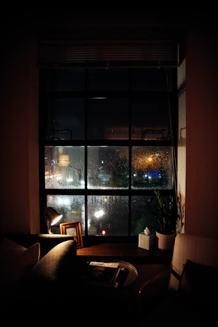 A cozy home window view showing rainy weather and a person smiling inside.
