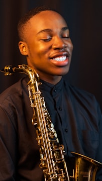A young man is joyfully holding a saxophone against a dark background. His eyes are closed, and he is smiling, conveying a sense of enjoyment and passion for music. He is wearing a dark shirt, and the shiny brass of the saxophone stands out prominently.