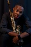A person sits with a saxophone held against their chest, wearing a dark long-sleeved shirt. The scene is dimly lit, highlighting the warm brass tones of the saxophone.