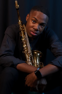 A person sits with a saxophone held against their chest, wearing a dark long-sleeved shirt. The scene is dimly lit, highlighting the warm brass tones of the saxophone.