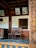 Wooden chairs are placed next to a window with bars under a wall adorned with framed pictures, including artwork of deities and an old family photograph. The setting includes wooden furniture and intricate carvings, suggesting a traditional or historical theme.