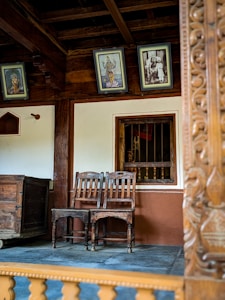 Wooden chairs are placed next to a window with bars under a wall adorned with framed pictures, including artwork of deities and an old family photograph. The setting includes wooden furniture and intricate carvings, suggesting a traditional or historical theme.