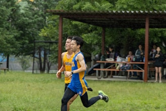 Two runners in athletic gear are competing on a grassy field, with spectators sitting on benches under a pavilion. The runners appear focused and determined as they move at a fast pace. Surrounding the area are lush green trees.