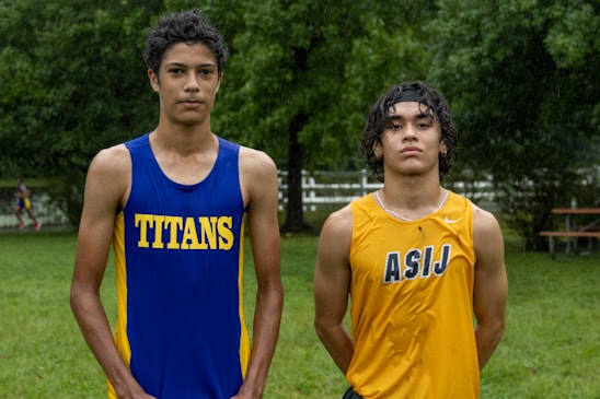 Two young athletes wearing sleeveless sports tops stand on a grassy field. The athlete on the left is wearing a blue top with the word 'TITANS' while the one on the right wears a yellow top with 'ASIJ'. The background features green trees and a white fence, with a bench visible on the right.