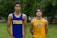 Two young athletes wearing sleeveless sports tops stand on a grassy field. The athlete on the left is wearing a blue top with the word 'TITANS' while the one on the right wears a yellow top with 'ASIJ'. The background features green trees and a white fence, with a bench visible on the right.